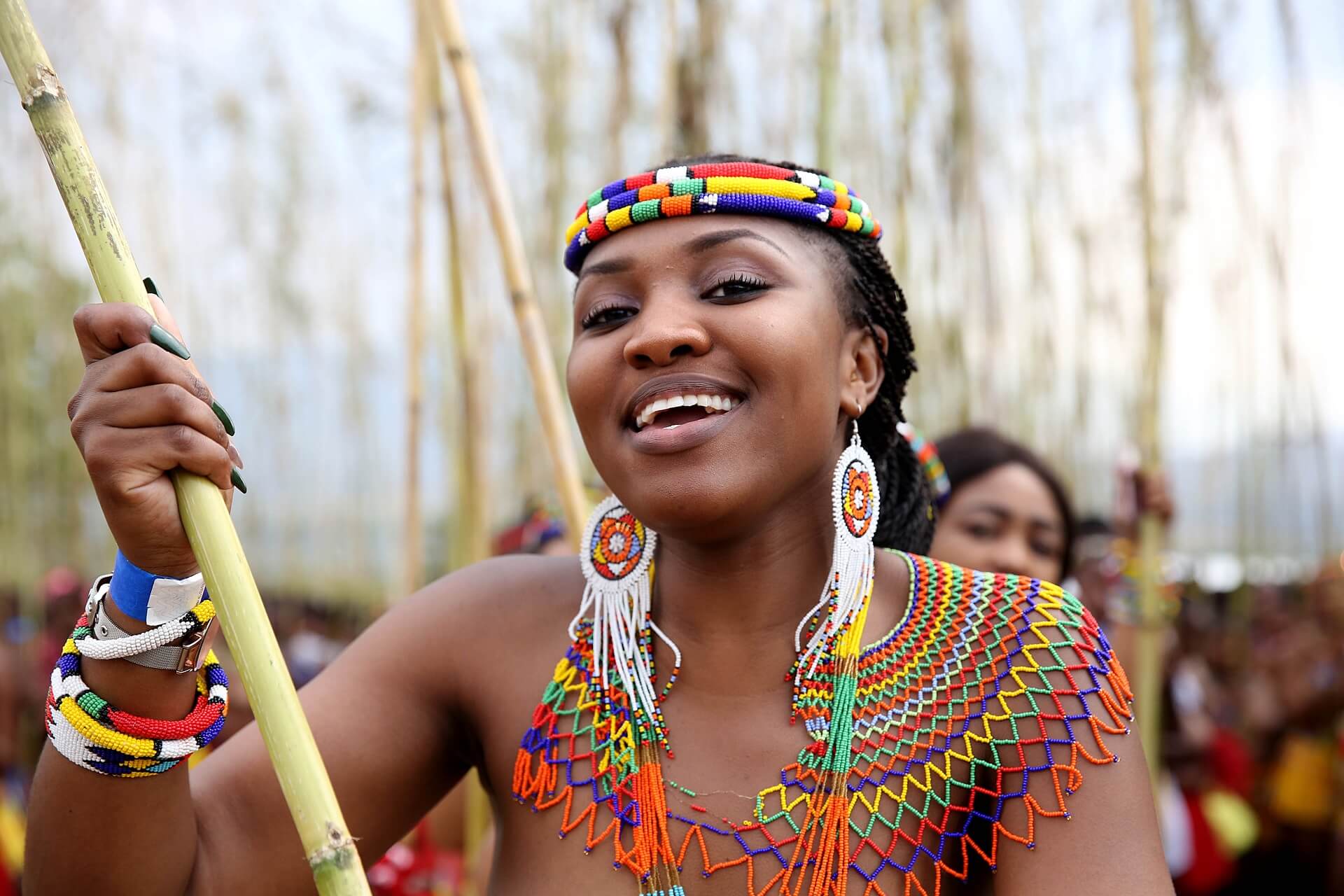 A person wearing colorful traditional African beads with Zulu clothing and beading smiles while holding a tall reed outdoors, offering a glimpse into the rich history and brand of South Africa—a perfect snapshot for anyone passionate about travel and tourism.