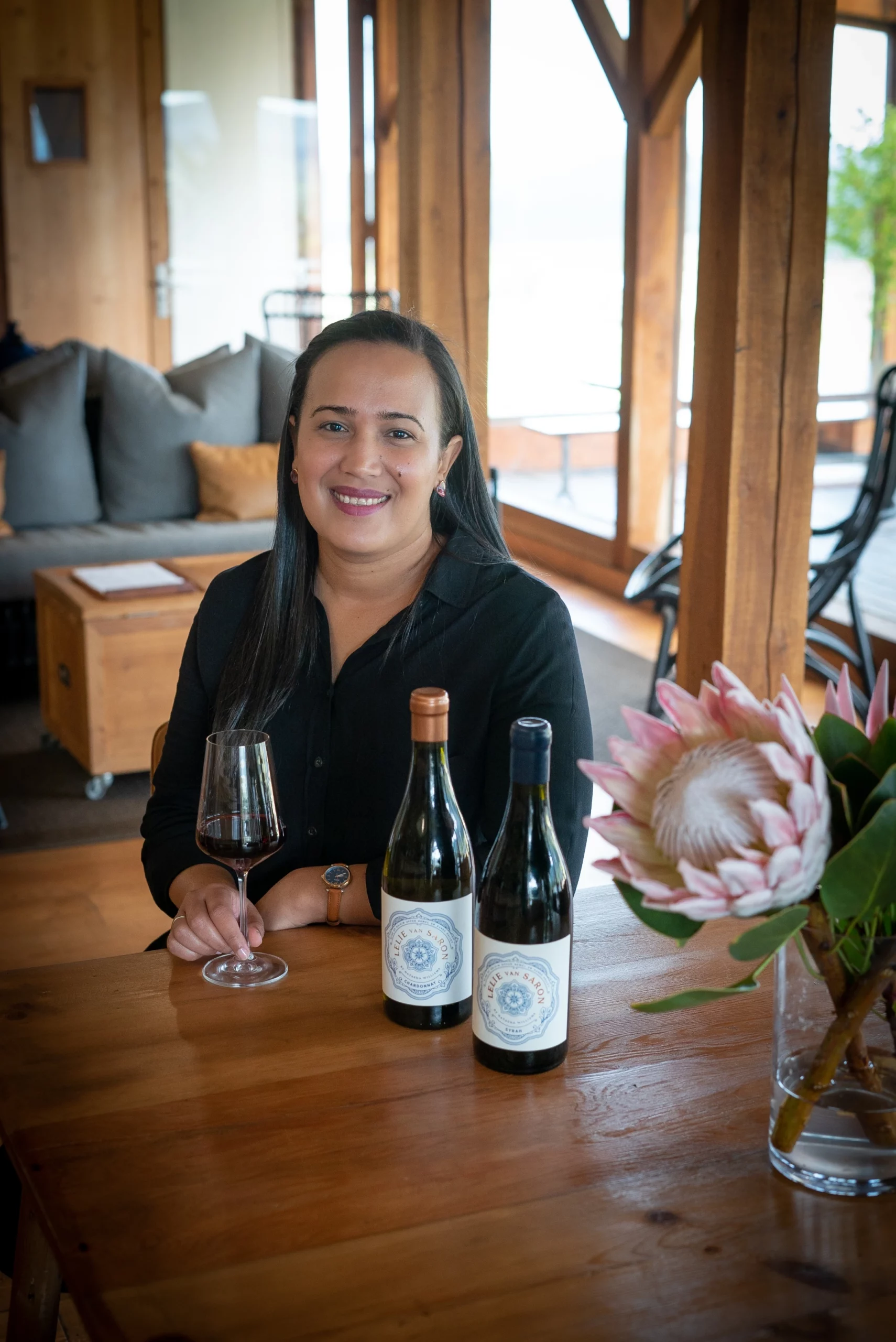Natasha Williams, winemaker, sitting at a wooden table with bottles of her Lelie van Saron wine made in the Cape. A glass of red wine and a protea flower are also present