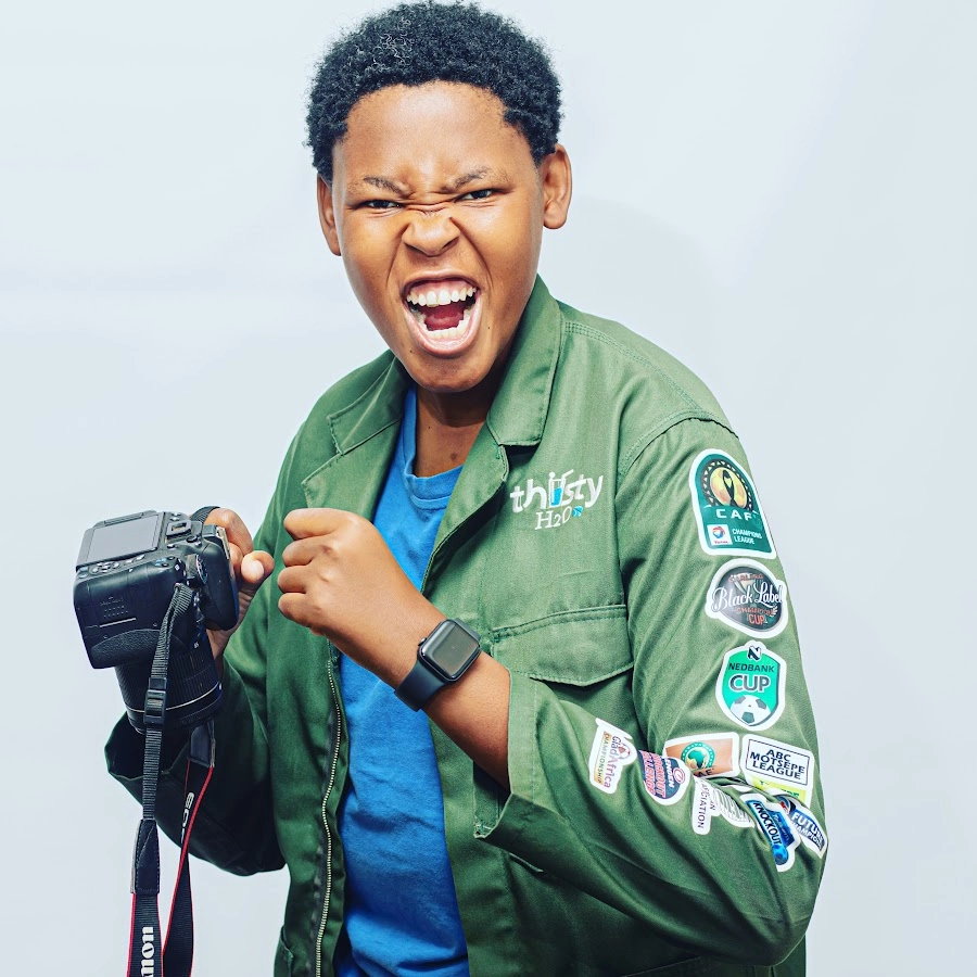 South African photographer, Pitsi Vincent Matlou—Vino Snap, wearing a green utility jacket adorned with various patches, holding a professional DSLR camera.